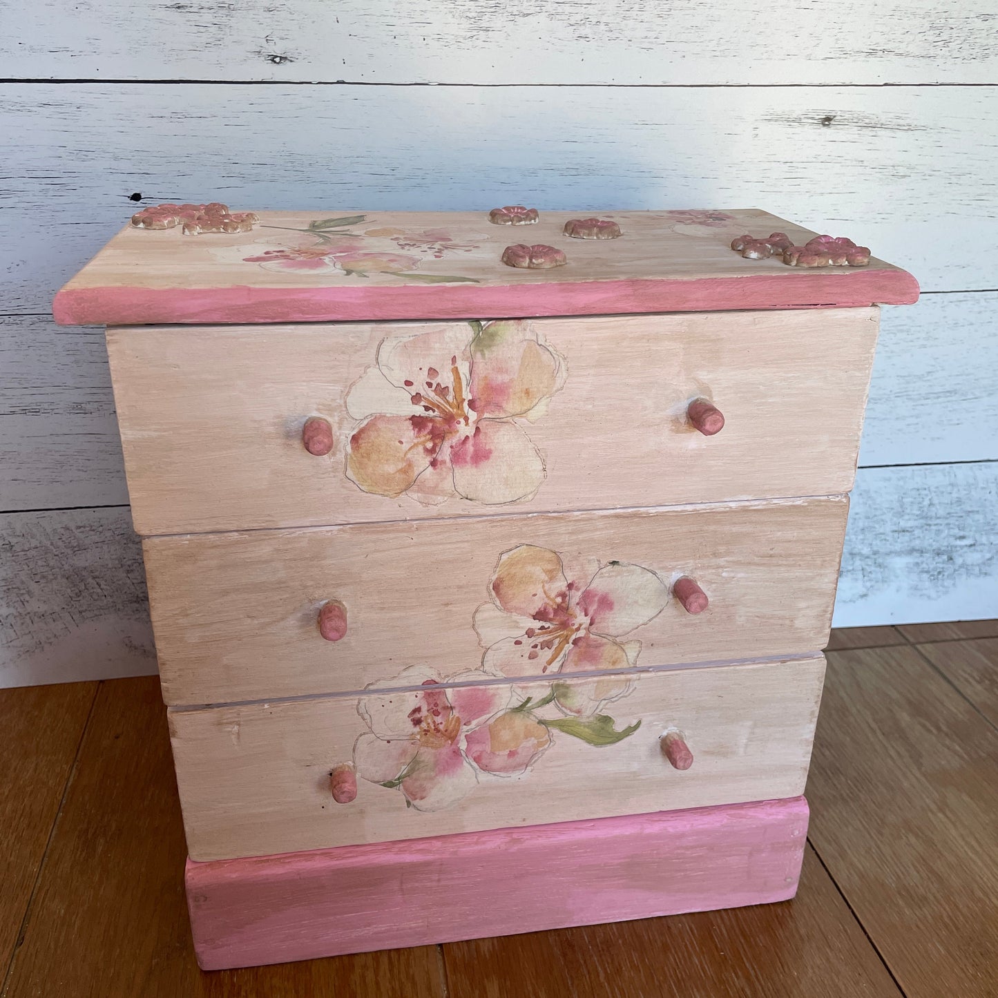Upcycled vintage wooden jewelry box with three drawers, hand-painted in soft blush and pink tones. Features delicate floral designs with watercolor-style blossoms on the front and top, along with rose-toned charm embellishments on the lid corners. The base and drawer knobs are painted in coordinating pink shades, creating a romantic, cottage-style look
