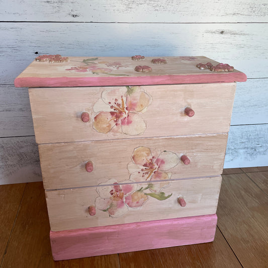 Upcycled vintage wooden jewelry box with three drawers, hand-painted in soft blush and pink tones. Features delicate floral designs with watercolor-style blossoms on the front and top, along with rose-toned charm embellishments on the lid corners. The base and drawer knobs are painted in coordinating pink shades, creating a romantic, cottage-style look