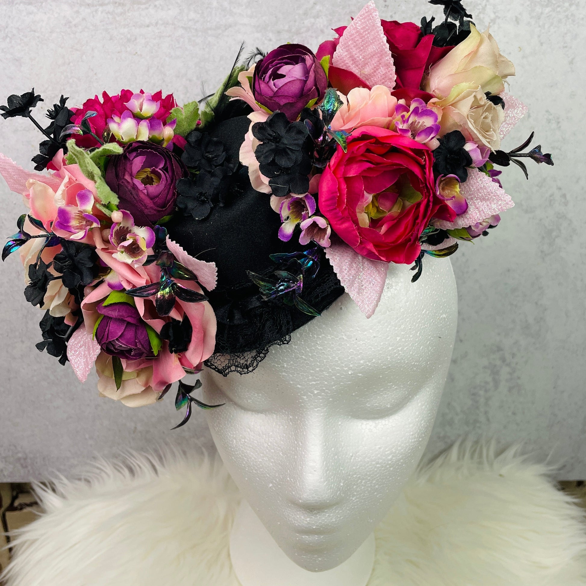 Floral mini top hat fascinator displayed on a mannequin head, featuring a dramatic arrangement of silk flowers in vibrant pink, purple, cream, and black tones with textured ribbons, lace, and iridescent accents—perfect for tea parties, vintage events, or whimsical photo shoots.