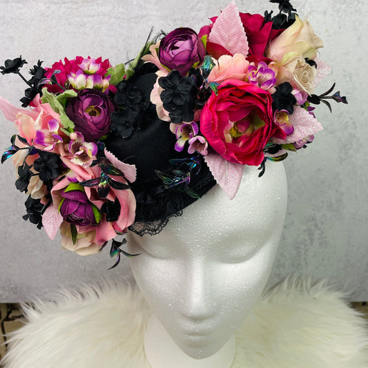 Floral mini top hat fascinator displayed on a mannequin head, featuring a dramatic arrangement of silk flowers in vibrant pink, purple, cream, and black tones with textured ribbons, lace, and iridescent accents—perfect for tea parties, vintage events, or whimsical photo shoots.