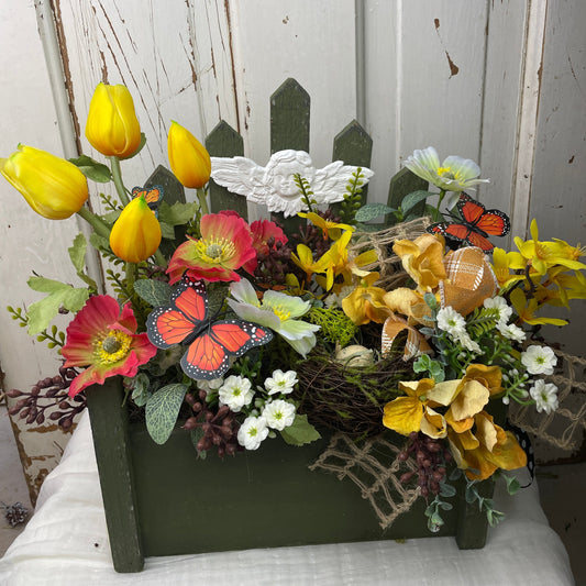 Faux spring floral arrangement in a green wooden picket fence box, featuring yellow tulips, red poppies, forsythia, white blossoms, butterflies, a grapevine nest with speckled eggs, and a cherub accent—perfect for cottage and garden-inspired decor.