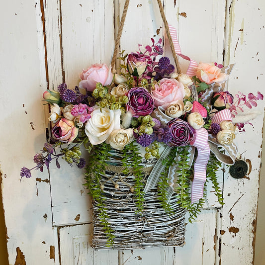 French cottage-style hanging basket filled with faux roses, ranunculus, and lavender in shades of pink, cream, and purple, accented with trailing greenery and layered ribbon bows, displayed on a chippy vintage white door.