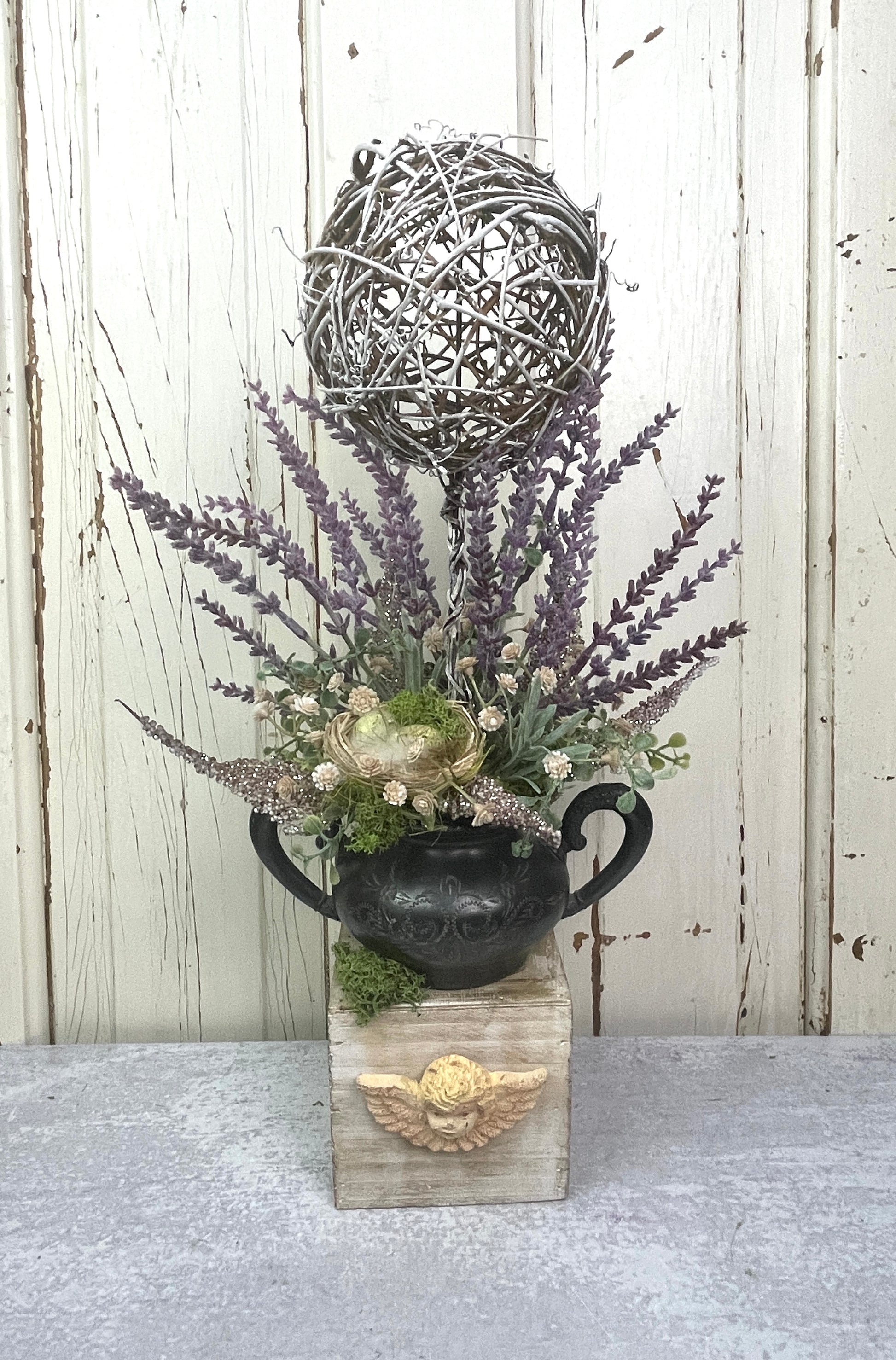 French cottage-style floral arrangement with faux lavender stems in a unique angel-embellished container, featuring a vintage silver sugar holder, grapevine topiary, and bird’s nest with feathers, styled in soft rustic tones.