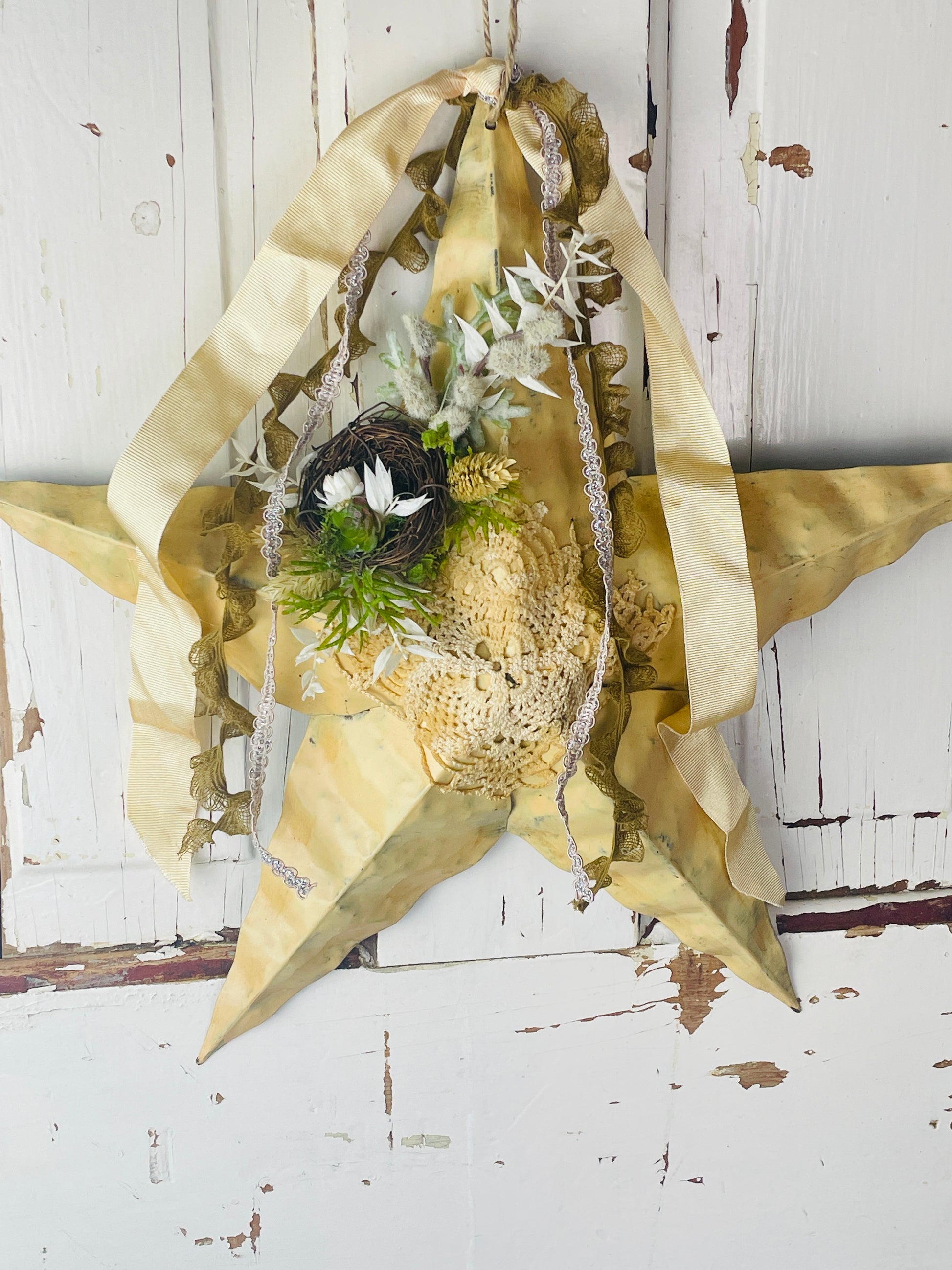 Primitive metal star wall hanging is yellow with dried floral accents, a small birds nest, and loose ribbons at the top