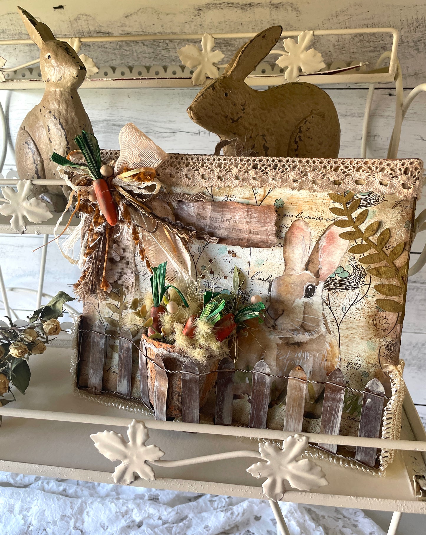 Vintage-style bunny-themed mixed media wall decor featuring a soft brown rabbit illustration, rustic wood picket fence, textured paper layers, lace trim, and a mini carrot bundle with greenery and fuzzy accents in a bark-style pot. Embellished with a neutral ribbon bow and displayed with distressed bunny figurines on a cream-colored metal shelf.