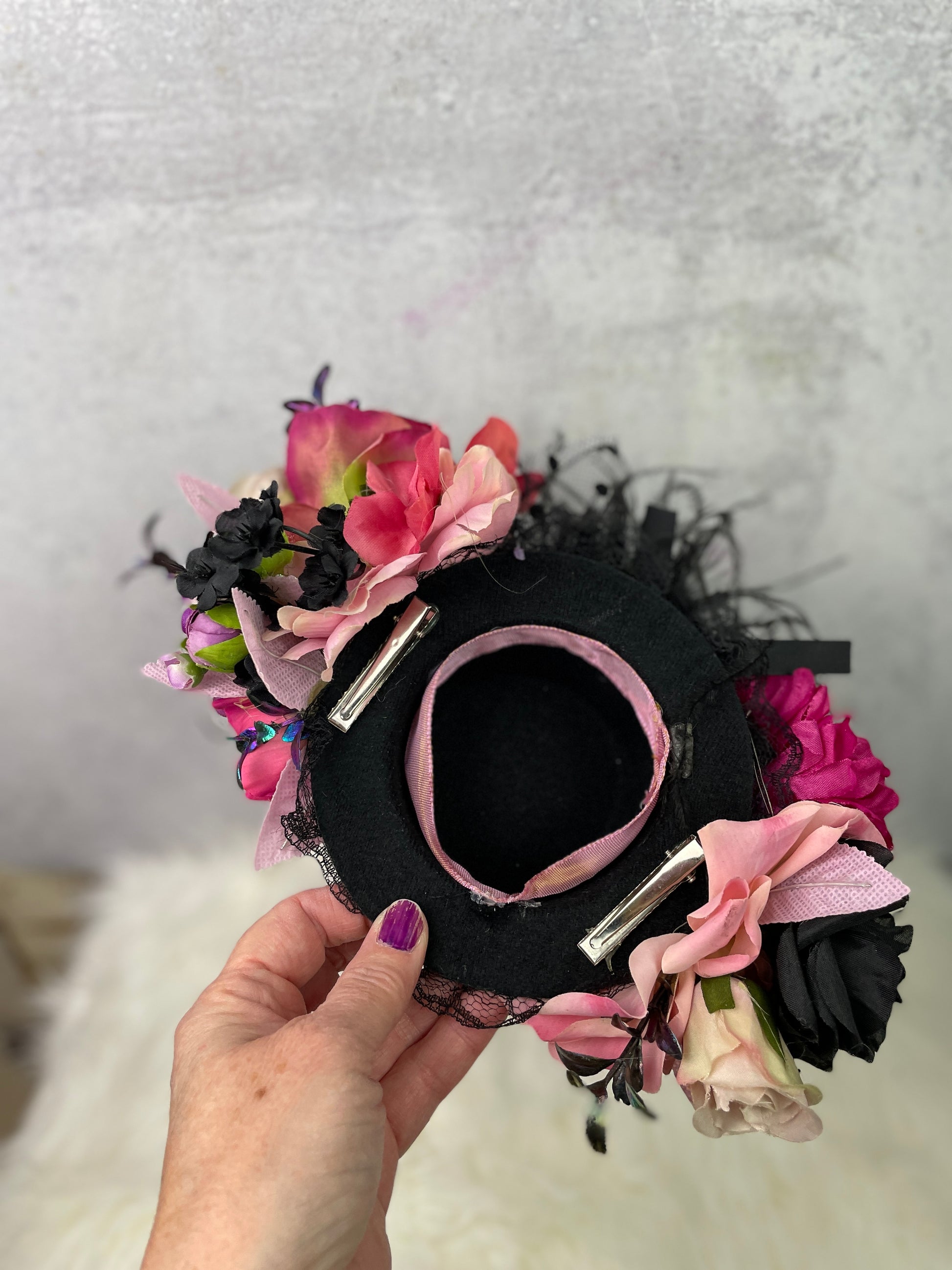 Hand holding a black mini top hat adorned with silk flowers in pink, purple, and black tones, shown from the underside with visible hair clips and pink ribbon lining for secure wear—perfect for vintage-inspired or whimsical styling.