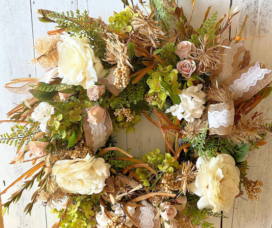 Handmade romantic wreath with cream roses, blush pink florals, greenery, burlap and lace ribbons, and rustic straw accents on a white wood background.