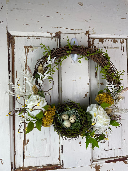 Handmade grapevine wreath featuring preserved white roses, yellow yarrow, dried florals, and a decorative bird nest with speckled eggs, perfect for cottagecore or French country door decor.