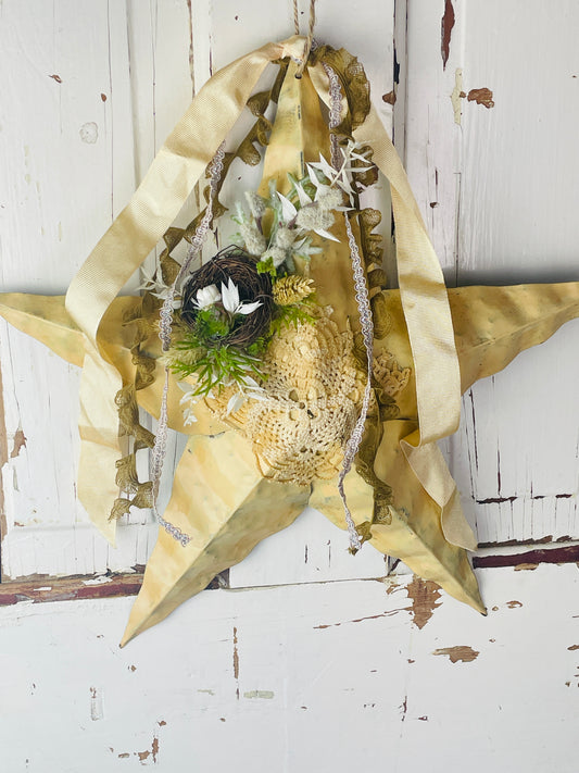 Primitive metal star wall hanging is yellow with dried floral accents, a small birds nest, and loose ribbons at the top 