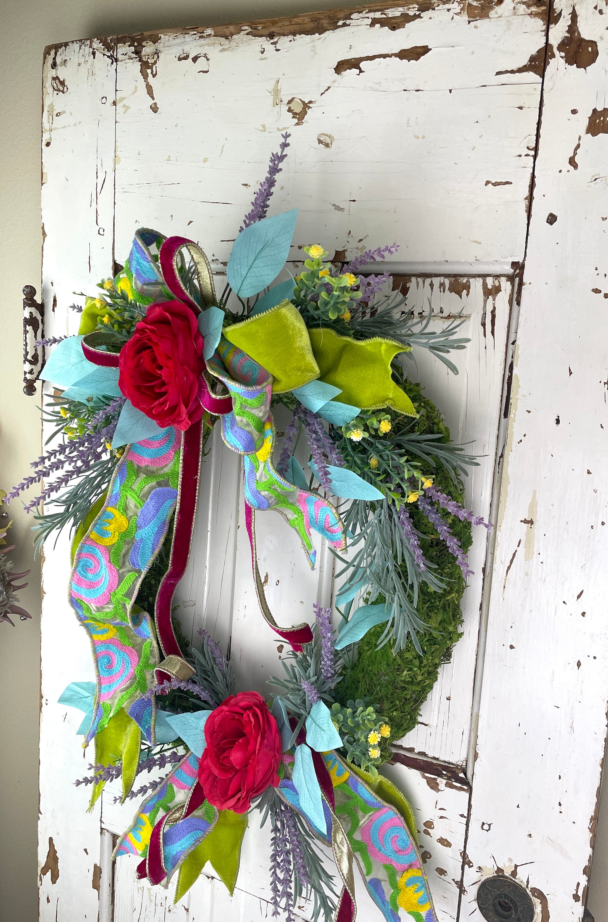 a moss oval wreath hangs on a vintage door with bright roses and velvet and tapestry ribbons hanging from t 